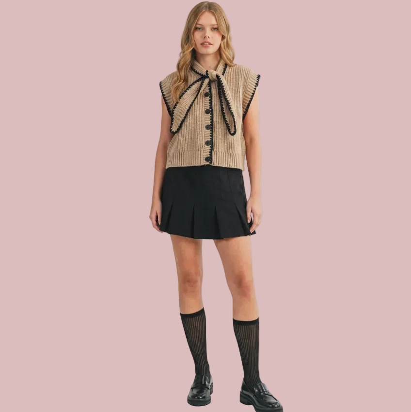 A model stands against a plain beige studio background wearing a sleeveless beige knit vest with black trim and a large bow detail, paired with a black pleated mini skirt, black knee-high socks, and black shoes.