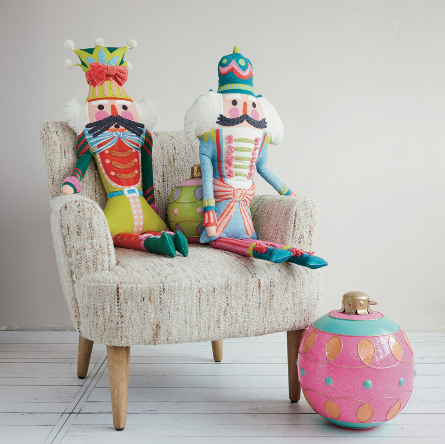 Two colorful fabric nutcracker dolls sit on a cream upholstered armchair on a light floor against a plain wall.