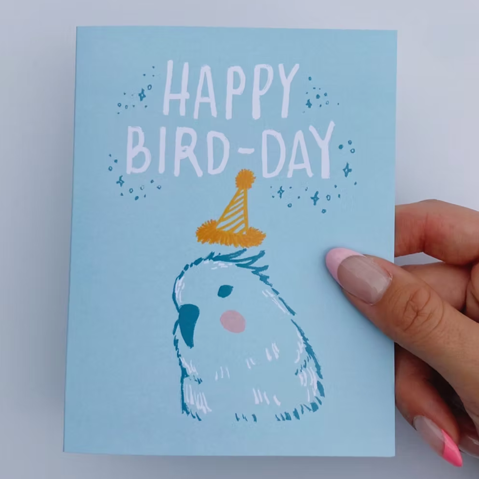 A blue greeting card that has a drawing of a bird wearing a birthday hat and the words, "Happy Bird-day" written above it.