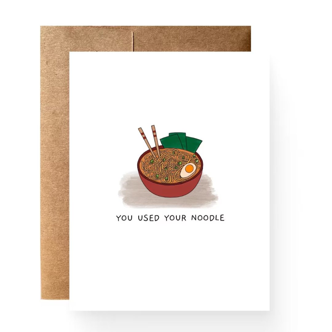 White greeting card with a watercolor painting of a maroon red bowl of ramen with lots of noodles, peas, and half of a boiled egg. Seaweed and chopsticks are sticking out of the bowl and the phrase, "You used your noodle", is in written font below. 