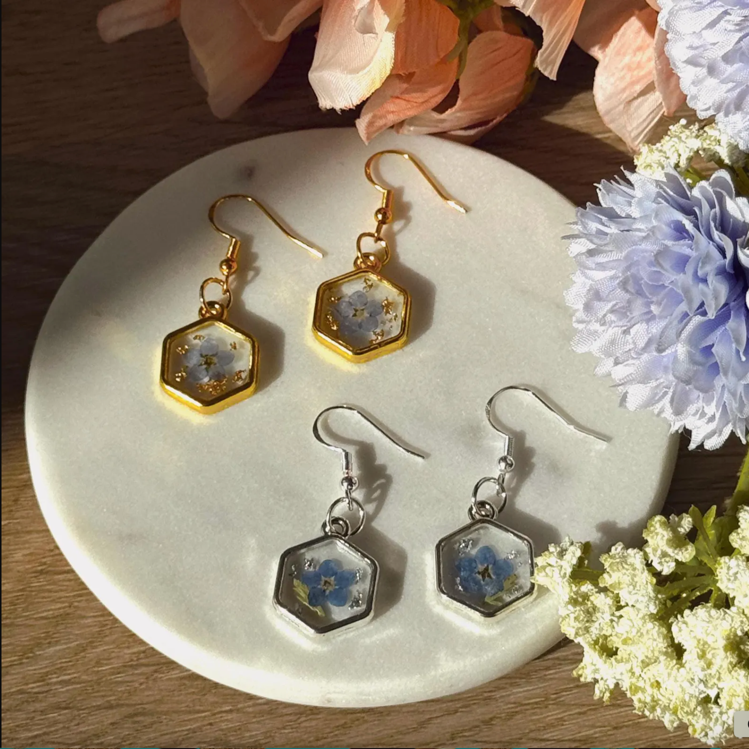 Dried Forget Me Nots placed in resin, surrounded by a hexagon 18k gold frame in a dangly earring. Displayed next to silver earrings of the same shape. 