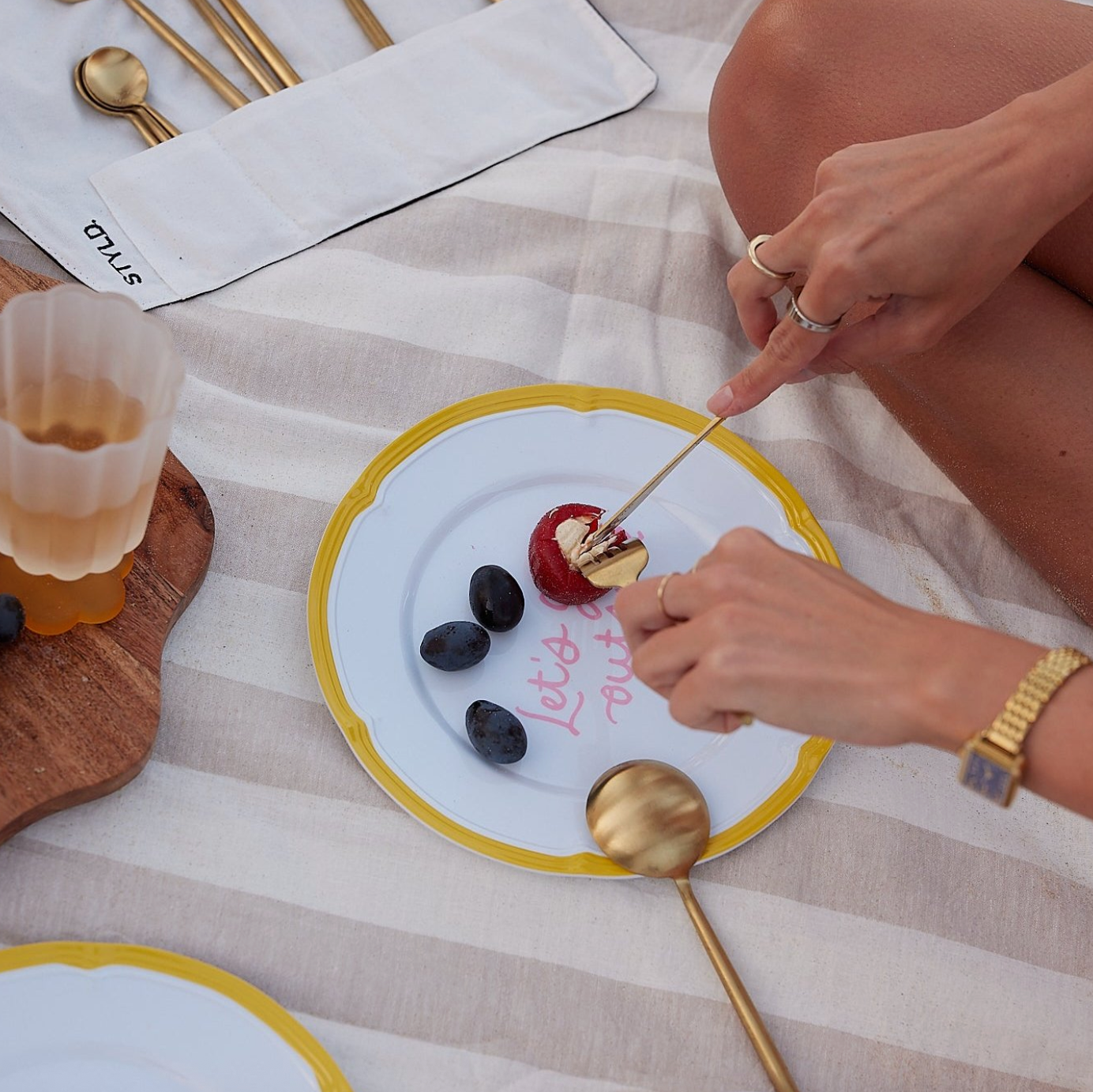 Two hands using gold utensils to cut into a dessert on a picnic blanket. 