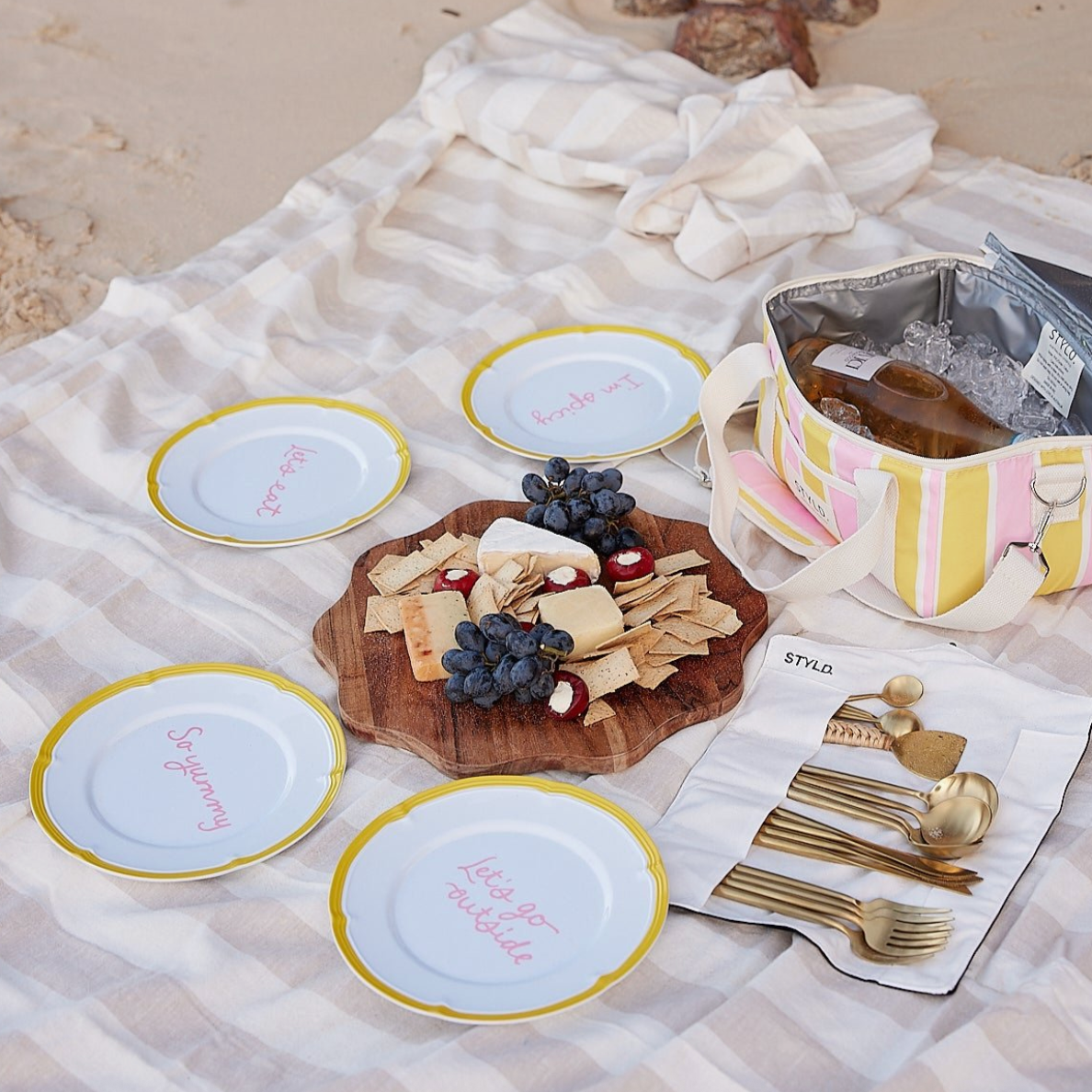 A picnic scene with four plates laid around a charcuterie board on a picnic blanket on the beach. 