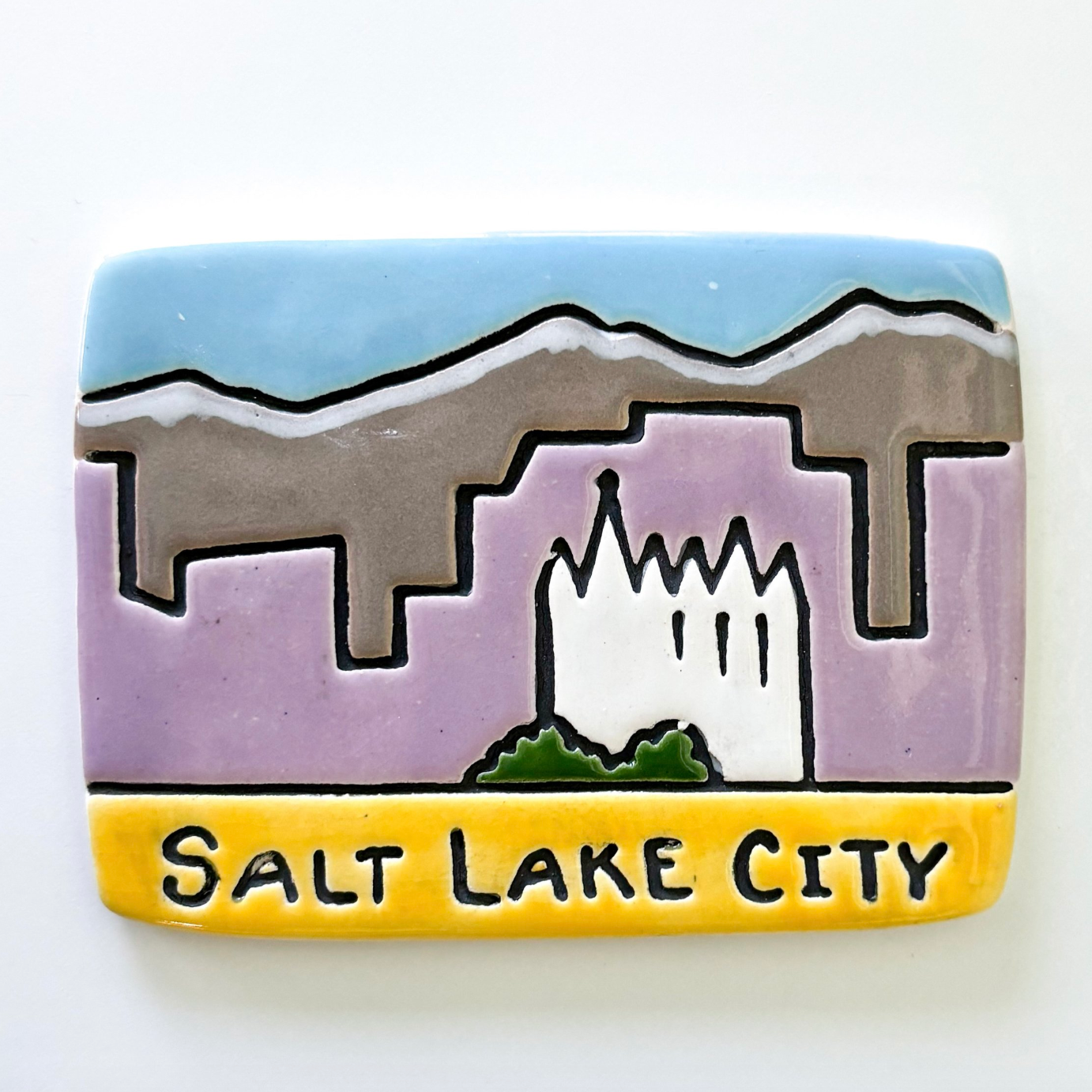 Ceramic ornament featuring a pared-down version of the SLC skyline. The temple is prominent with purple buildings behind it, grey, snow-capped mountains, and a blue sky. 