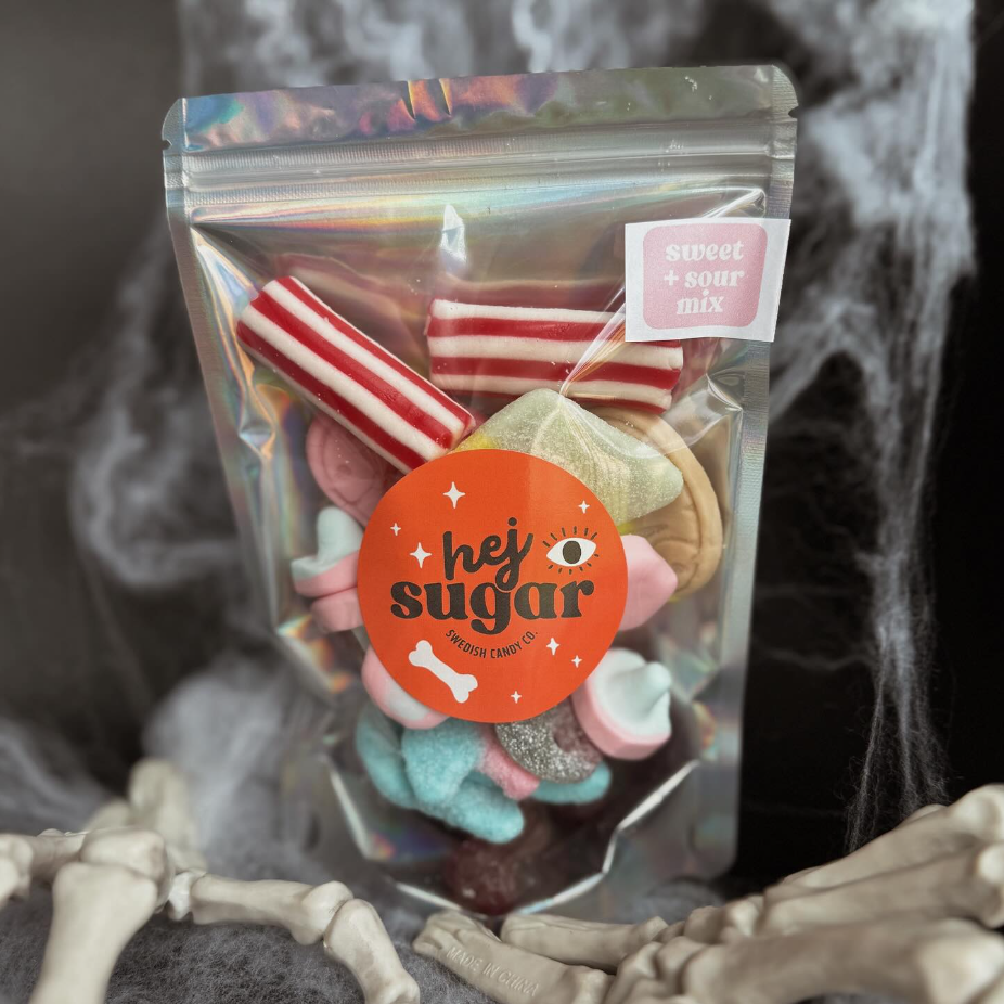 A holographic bag labeled “hej sugar” filled with colorful Swedish candies in various shapes, set against a dark background with fake spider webs and plastic skeleton decorations. The bag also has a pink sticker that reads “sweet + sour mix.”
