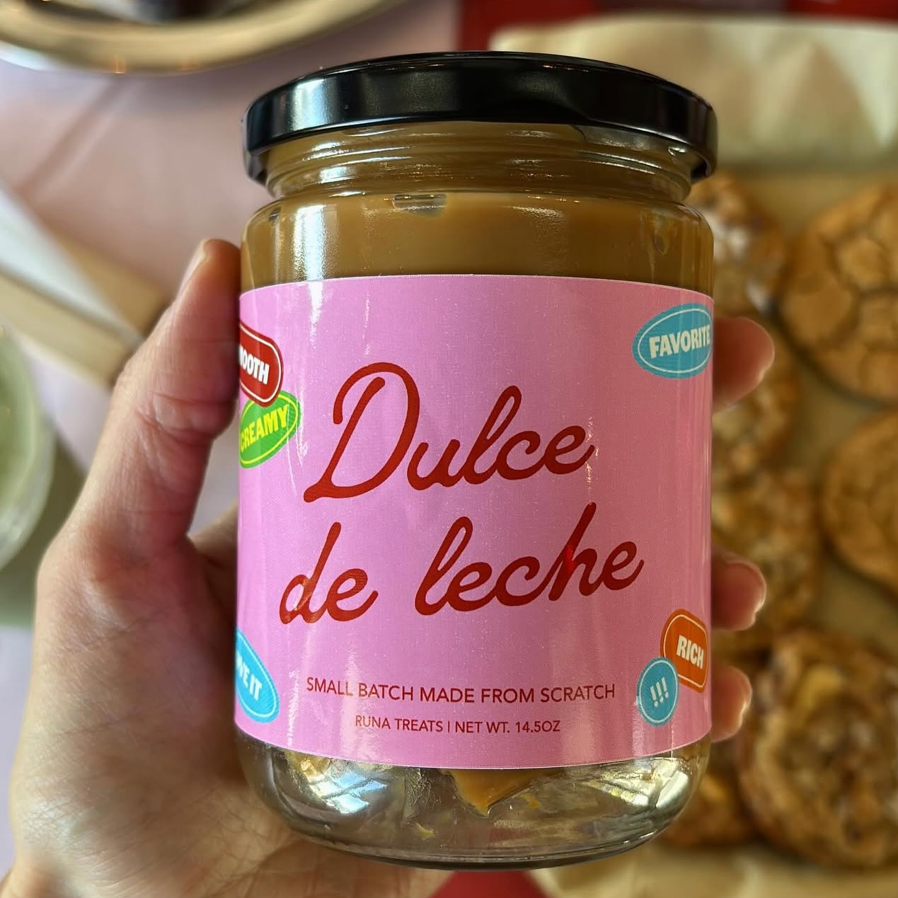 A hand holding a jar of Runa Treats Dulce de Leche with a pink label that reads “Dulce de leche” and “Small batch made from scratch.” The jar is filled with caramel-colored spread, and cookies are visible in the background.