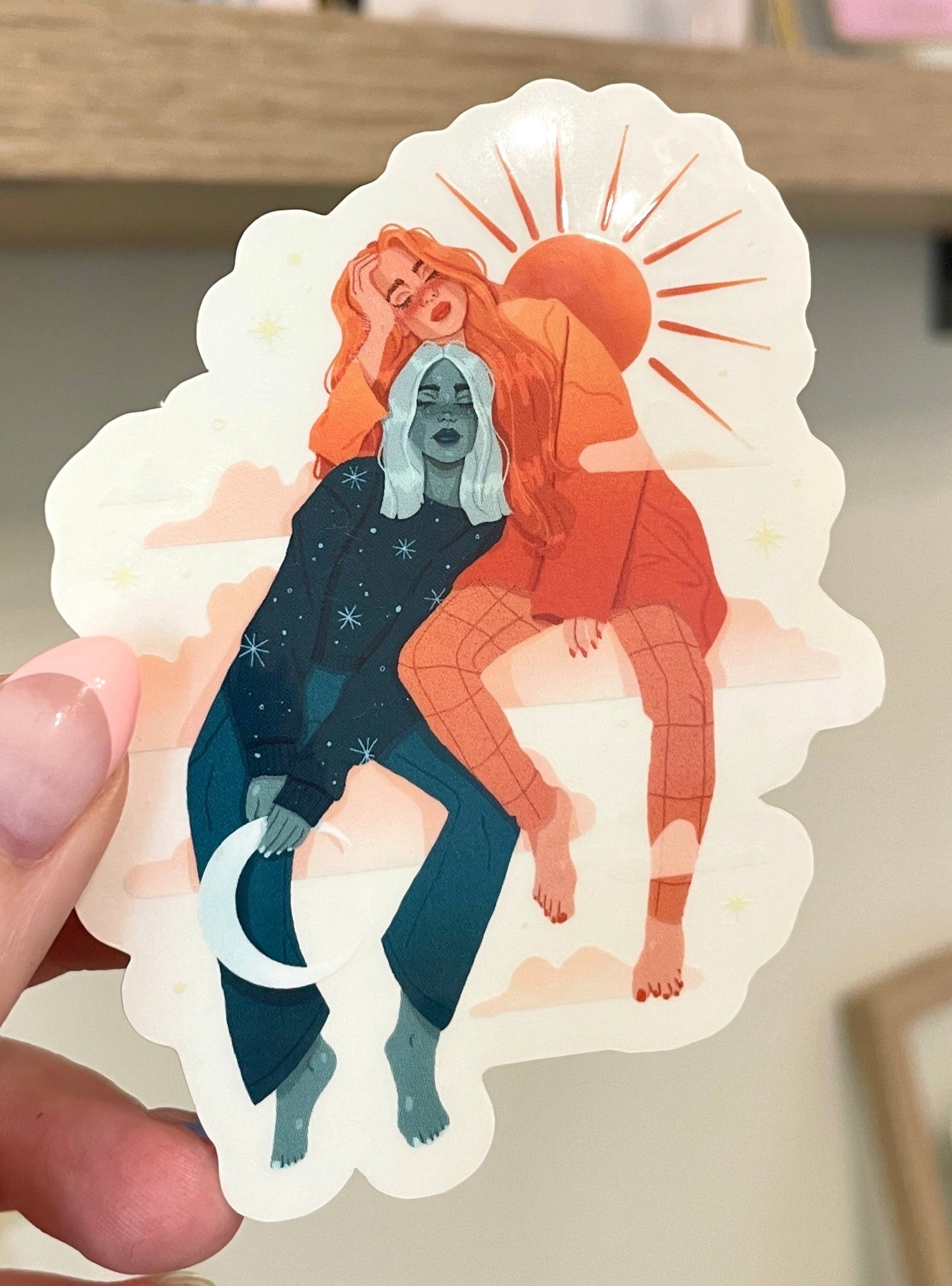 Sky sisters sticker with clear back. One sister is in a blue-green hue. She has shoulder length blonde hair and is holding a crescent moon. She is leaning against her sister who is in an orange hue. The orange sister has the sun behind her and is resting her chin on her sister's head. They are sitting on clouds together. 