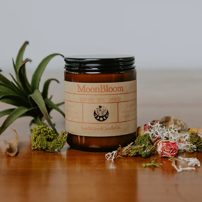 A soy wax candle named 'Moonbloom' in an amber jar with a label, surrounded by decorative dried plants and flowers on a wooden surface.