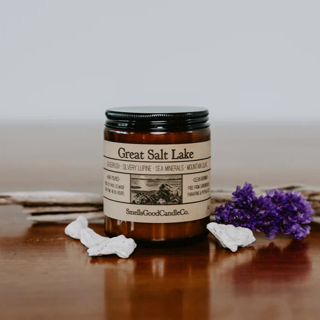 A soy wax candle jar with a label reading 'Great Salt Lake' on a wooden surface, flanked by a sprig of dried plant and a small purple flower.
