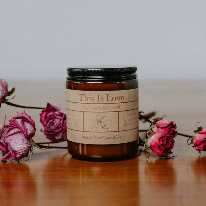 A brown glass jar scented candle labeled 'This Is Love' with dried pink roses lying in front of it, placed on a wooden surface.