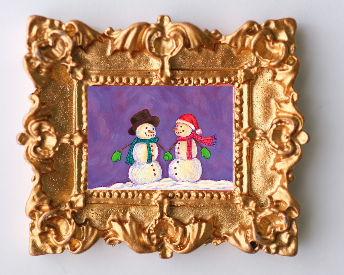 Ornate gold-framed Christmas magnet featuring two cheerful snowmen wearing colorful scarves, mittens, and winter hats against a purple background, with a snowy ground and vintage-inspired decorative frame.
