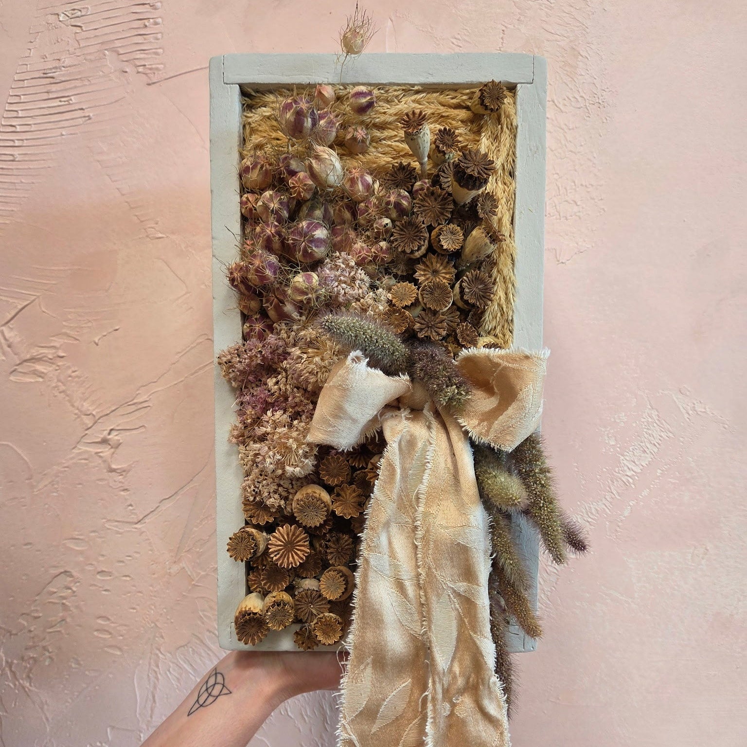 An arrangement of dried flora and fauna in  a light gray 8 x 14 wooden frame. Hues of gold, pink, and light green fill the space, and there is a cream colored ribbon beautifully adorned in the lower right quadrant. 