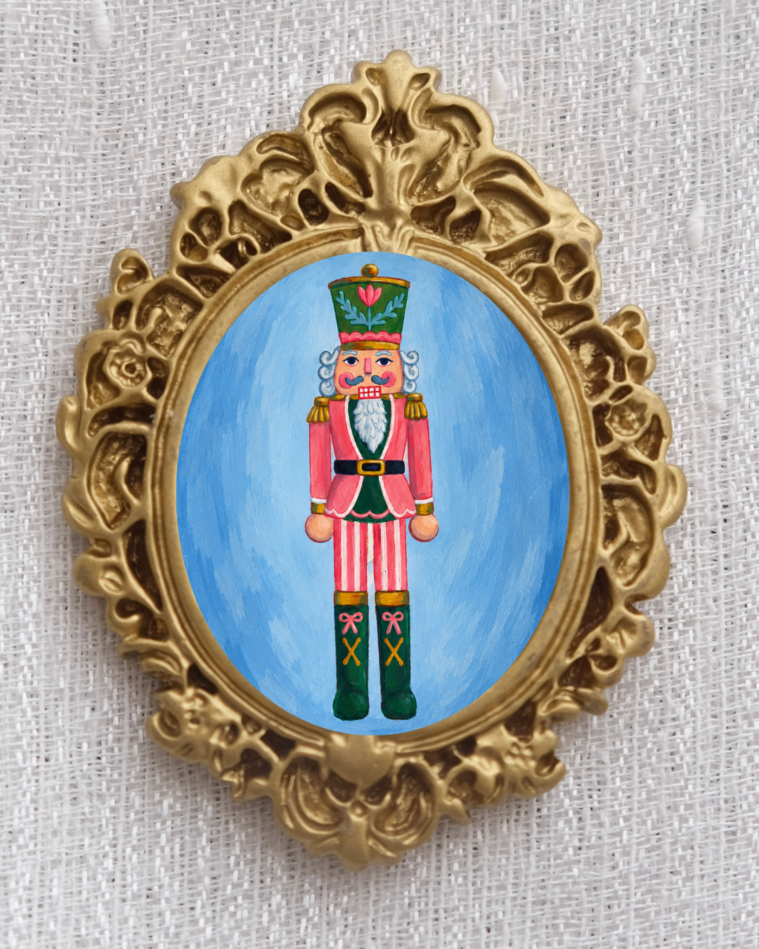 Ornate gold-framed Christmas magnet featuring a colorful Nutcracker soldier in a red, green, and gold uniform against a blue background, with an intricate vintage-inspired decorative frame.