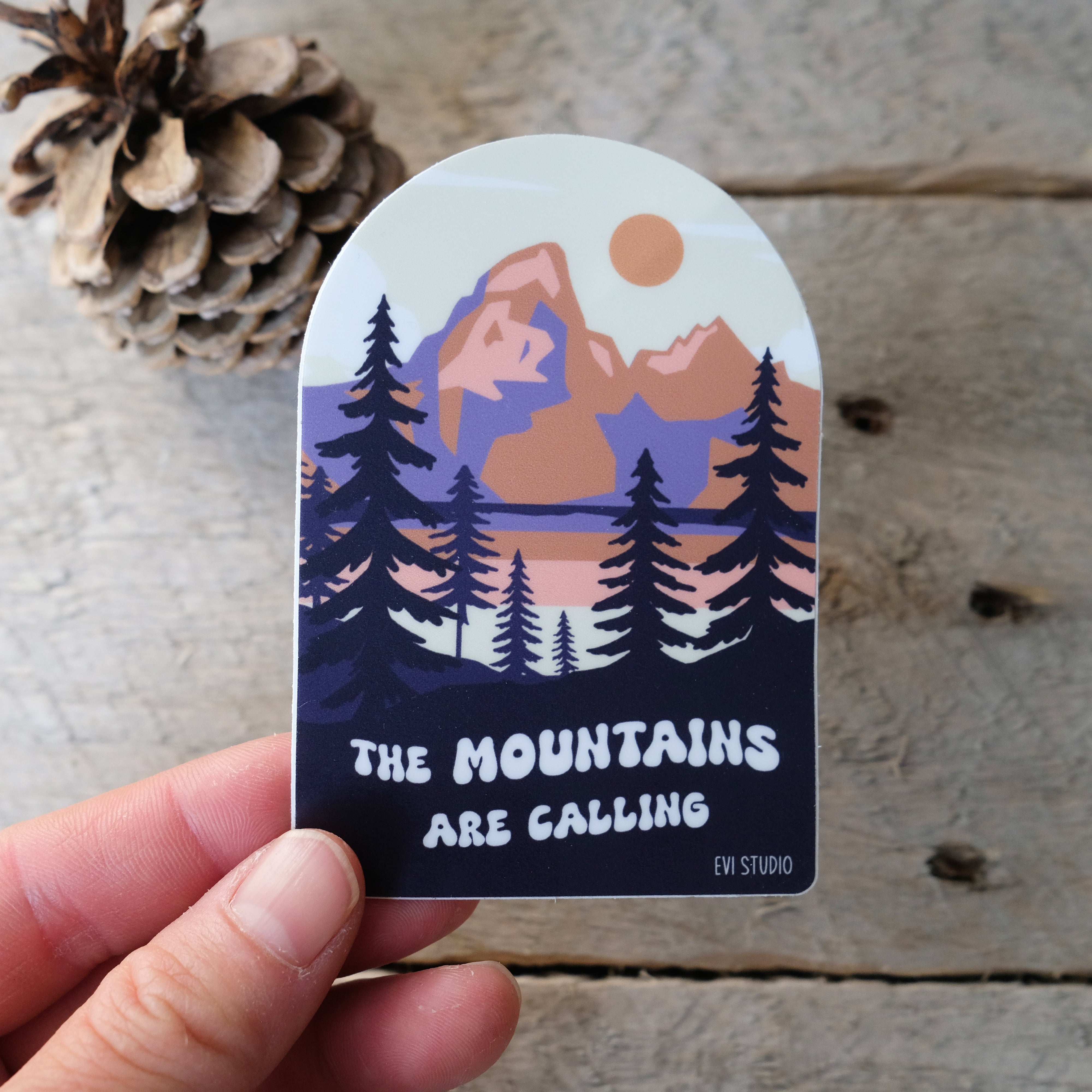 A hand holds a sticker with an illustration of mountains, pine trees, and a setting sun in muted orange, purple, and cream tones. The sticker reads “The mountains are calling” with “Evi Studio” printed below. A pinecone and rustic wooden surface are in the background.