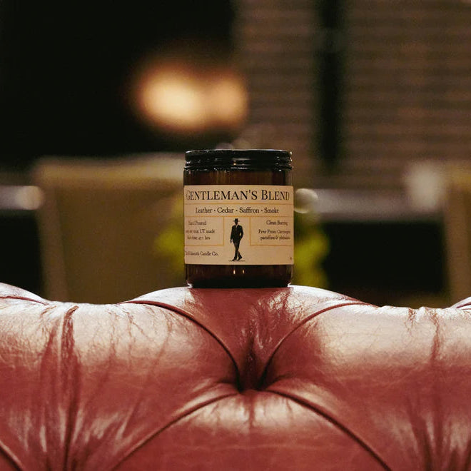 This image features the **Gentleman's Blend** candle once again, but now it's styled in a more luxurious and cozy setting. The candle sits on the backrest of a deep red tufted leather sofa, with a softly lit, upscale background that includes plush chairs and ambient lighting. The setting emphasizes a masculine, refined atmosphere—matching the scent notes of **leather, cedar, saffron, and smoke**.