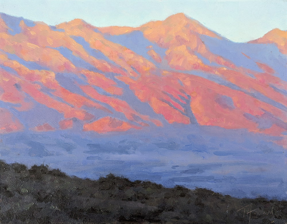 The Lone Peak mountain peak at dusk. The mountain is blue and purple with a blue sky in the background. In the foreground is greenery. 