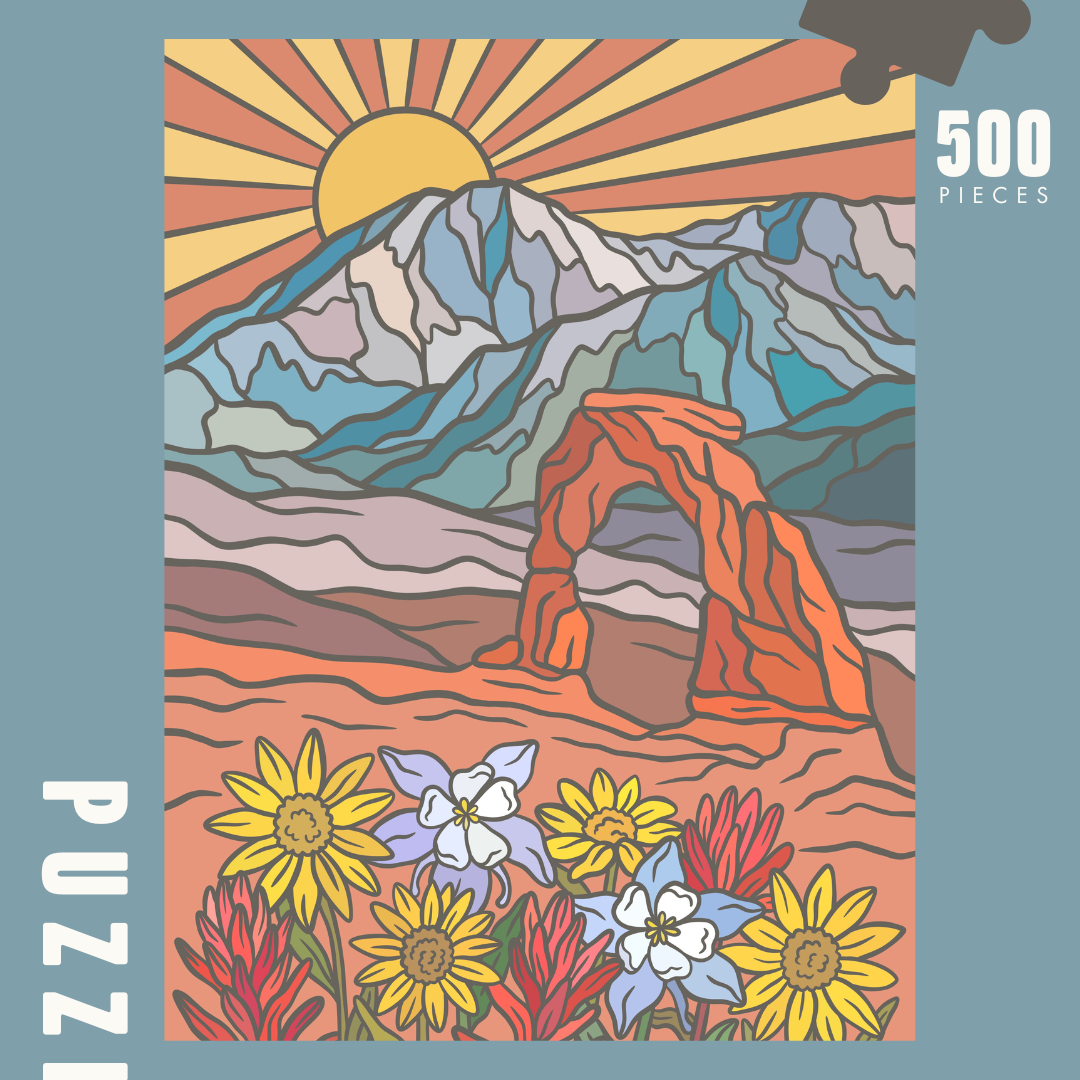 Image of puzzle box, showing full image of puzzle, Design includes a mountain range in the back in shades of blue, the iconic delicate arch, and utah wildflowers in the forefront, all against a sunset. 