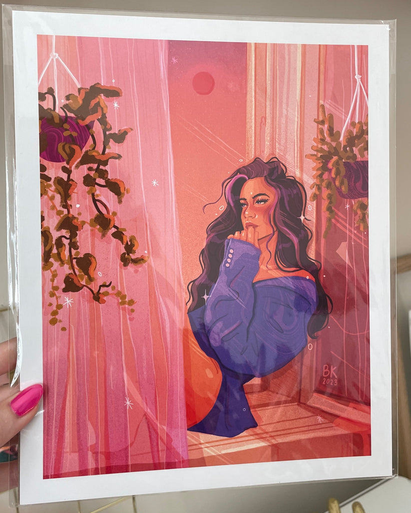 An 8x10 print of a woman sitting in a windowsill. The print is in pink hues and the woman is wearing an oversized blue sweater. She is surrounded by curtains and hanging plants. 