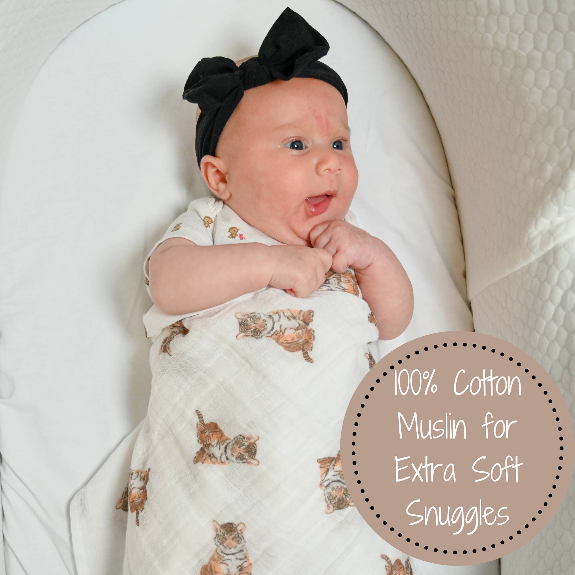Baby wrapped in a white swaddle with animal patterns, wearing a black headband, sitting in a white chair.