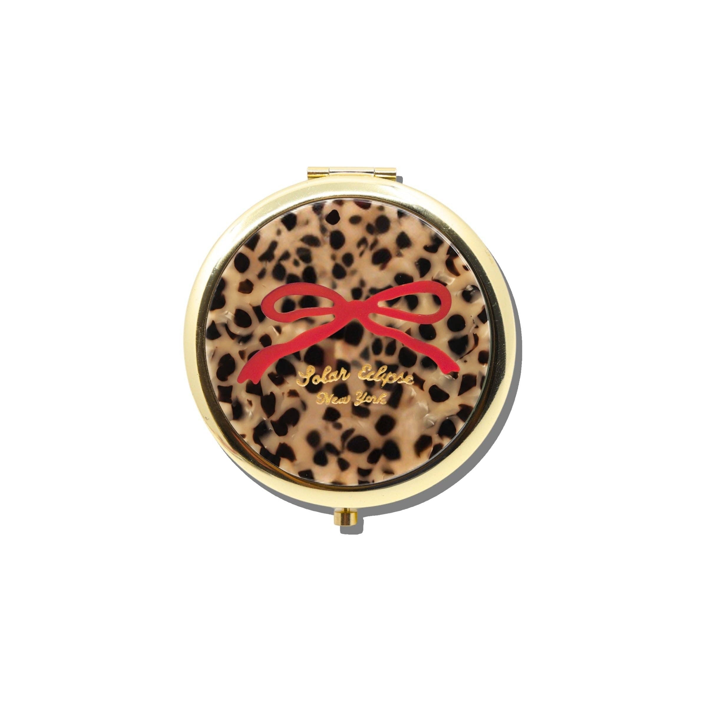 A round compact mirror with a leopard print pattern cover featuring a red bow graphic and gold lettering, framed in gold-toned metal, set against a plain white background.