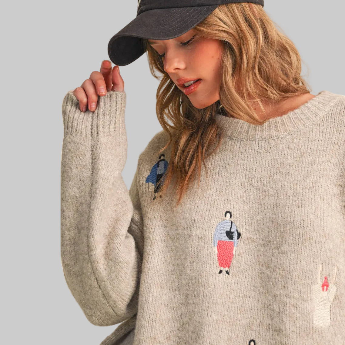 A woman wearing a light grey knit sweater with small colorful women dressed in fun clothing patches, paired with gray polka-dot pants, against a plain beige studio background.