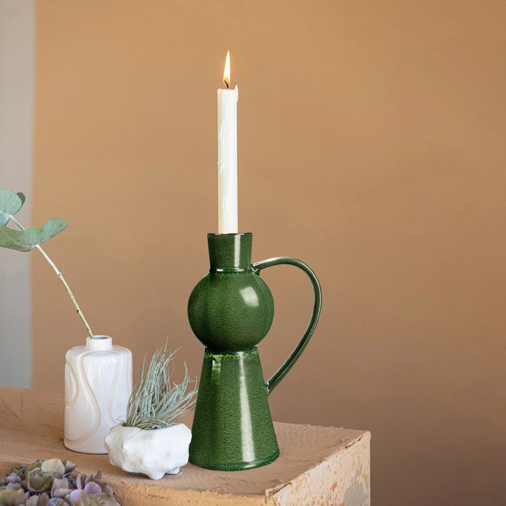 Illuminate the home with this green stoneware taper holder with a handle. This candle holder features a stoneware construction that has a reactive glaze that creates a beautiful variation of green hues. The candle holder has a unique shape that resembles a jug with a handle, adding some personality and flair to any space.