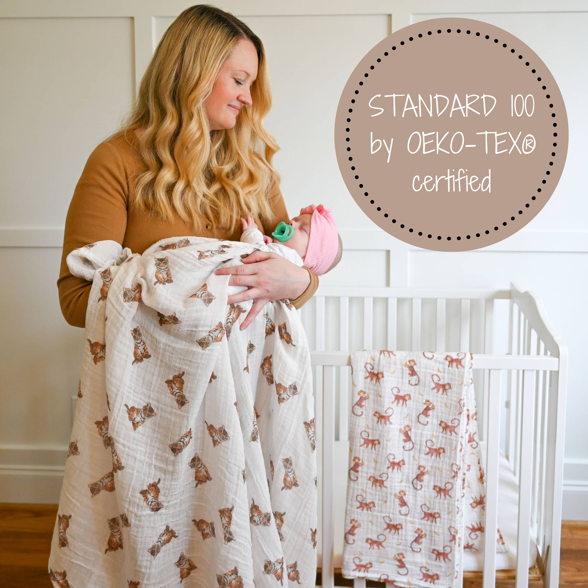 Woman holding a baby wrapped in a patterned blanket with a crib in the background, featuring OEKO-TEX certification.