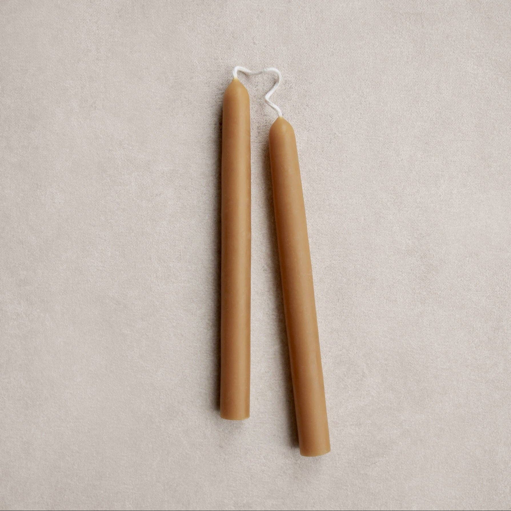 Two unpainted taper candles in a warm ochre color, standing upright on a textured beige surface.