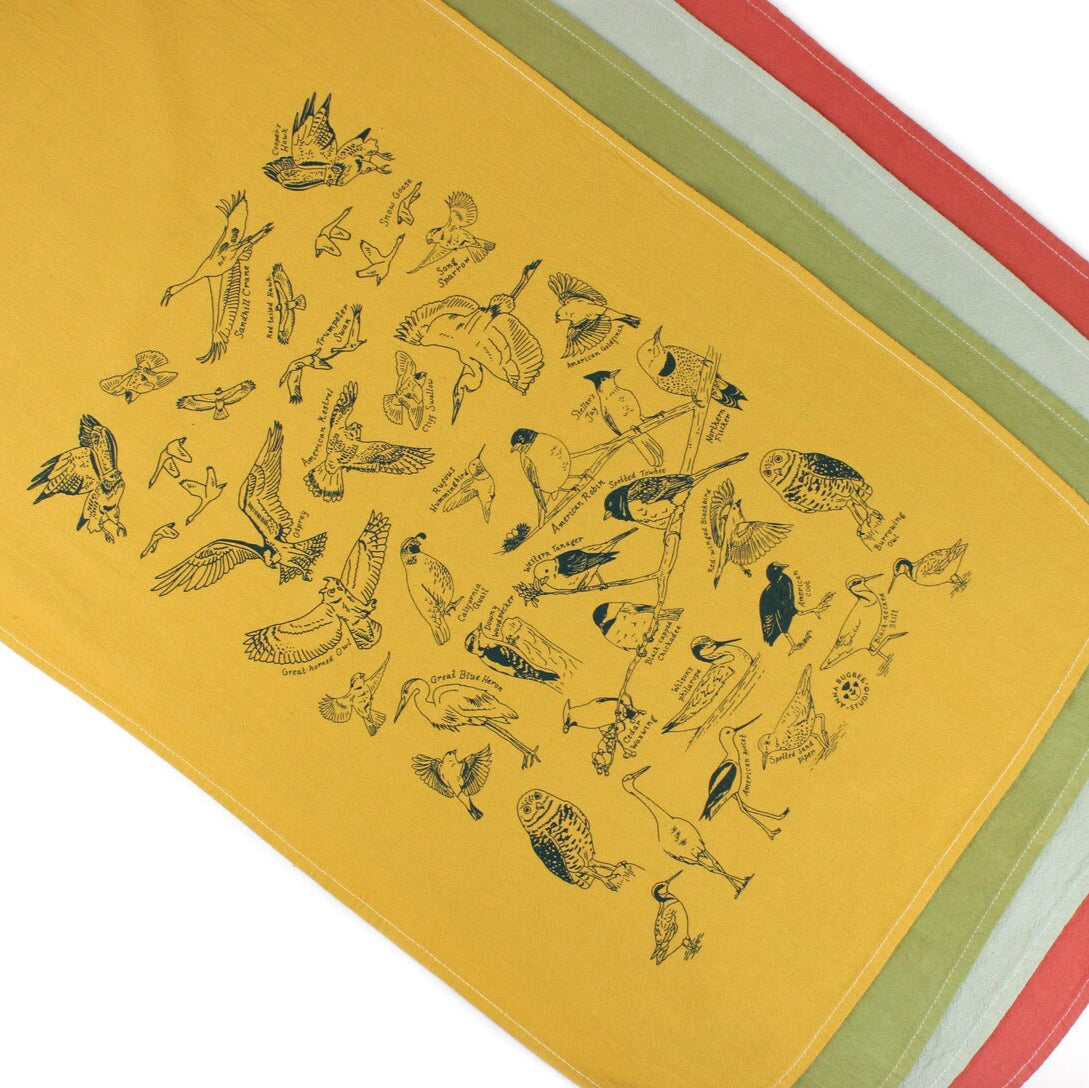 This image shows four neatly stacked cloth napkins or tea towels in warm tones—mustard yellow, olive green, pale blue, and burnt orange. The top towel features a detailed print of various bird species, each carefully labeled, creating a charming, nature-inspired design.