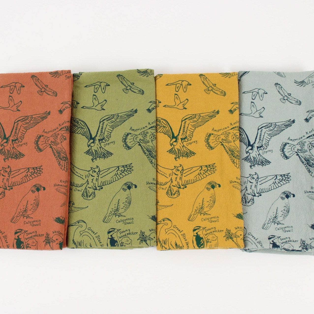 This image shows four neatly folded cloth napkins in a row, each in a different earthy color: rust orange, olive green, mustard yellow, and pale blue. Printed on each napkin are intricate line drawings of birds, labeled with their names—like “Osprey,” “American Kestrel,” and “California Quail”—creating a charming, nature-themed set.