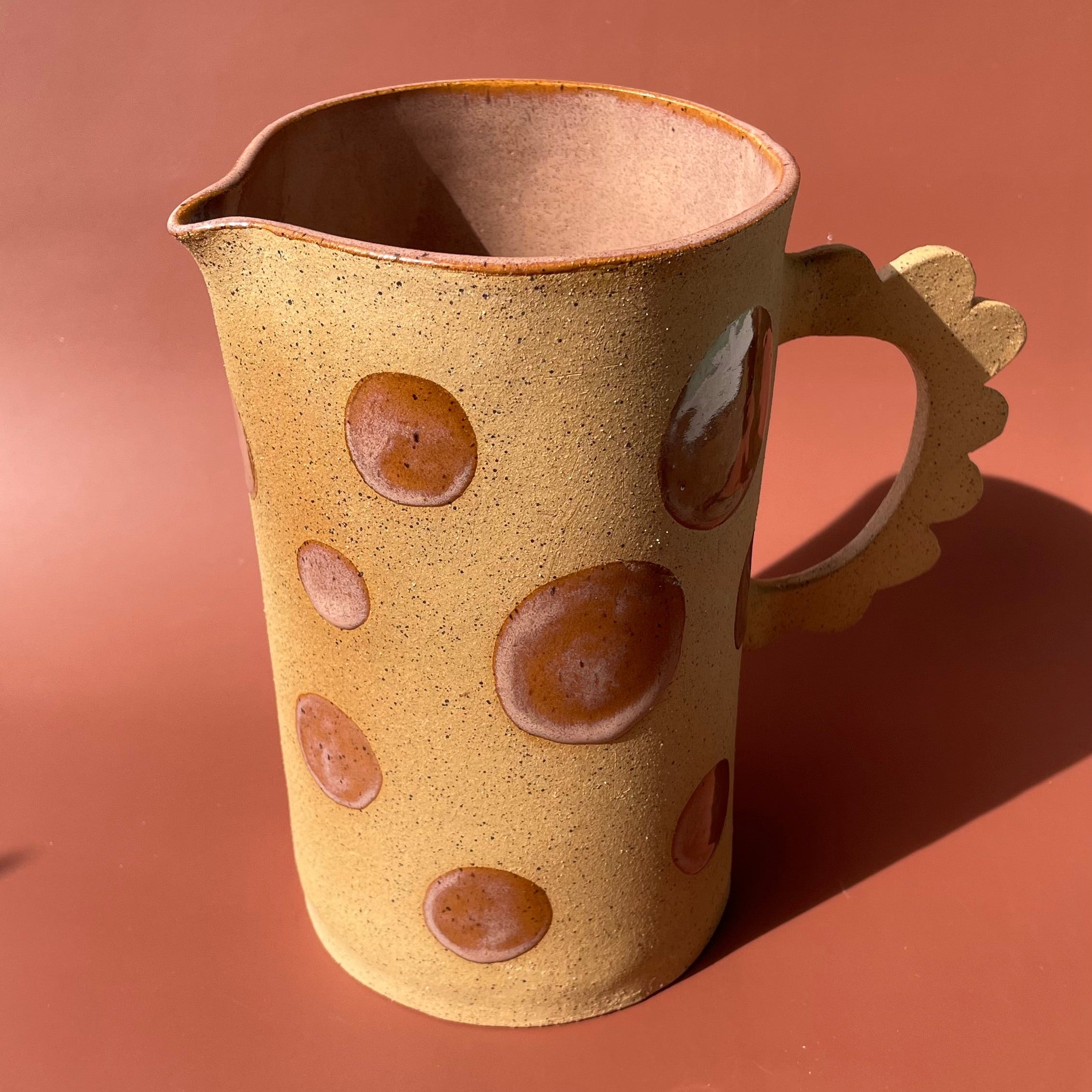 Handmade ceramic pitcher with a speckled brown finish, circular glaze accents, and a scalloped handle