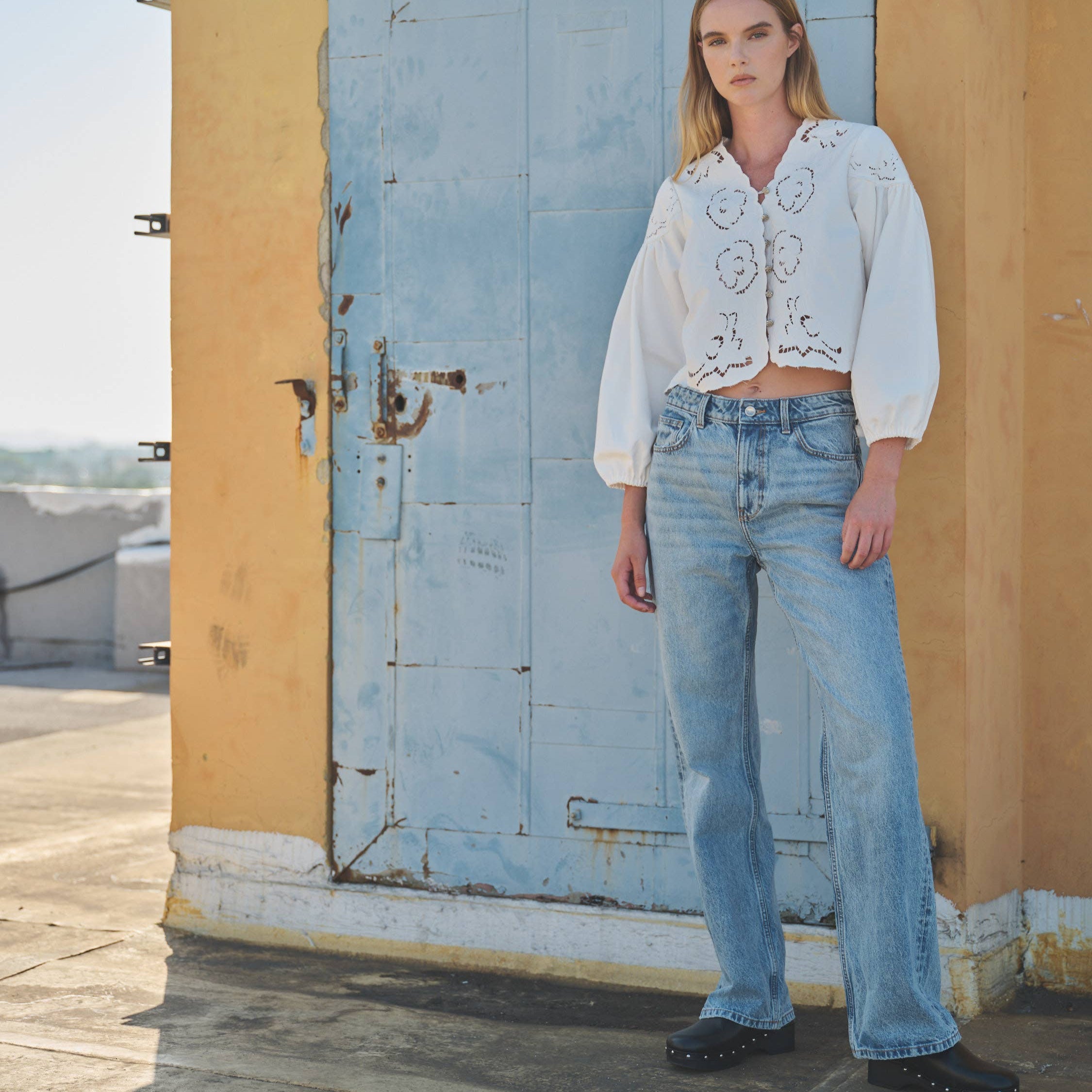 Woman wearing a white embroidered blouse and blue jeans standing in front of a rustic door.