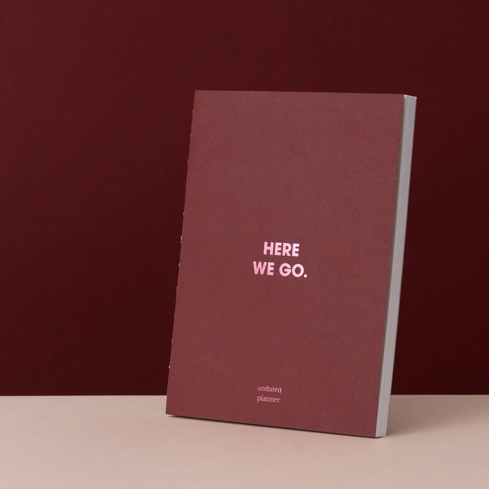 A hardcover notebook with a dark burgundy cover and light pink text reading 'HERE WE GO.' and 'undated planner,' standing upright on a beige surface against a matching dark burgundy background.