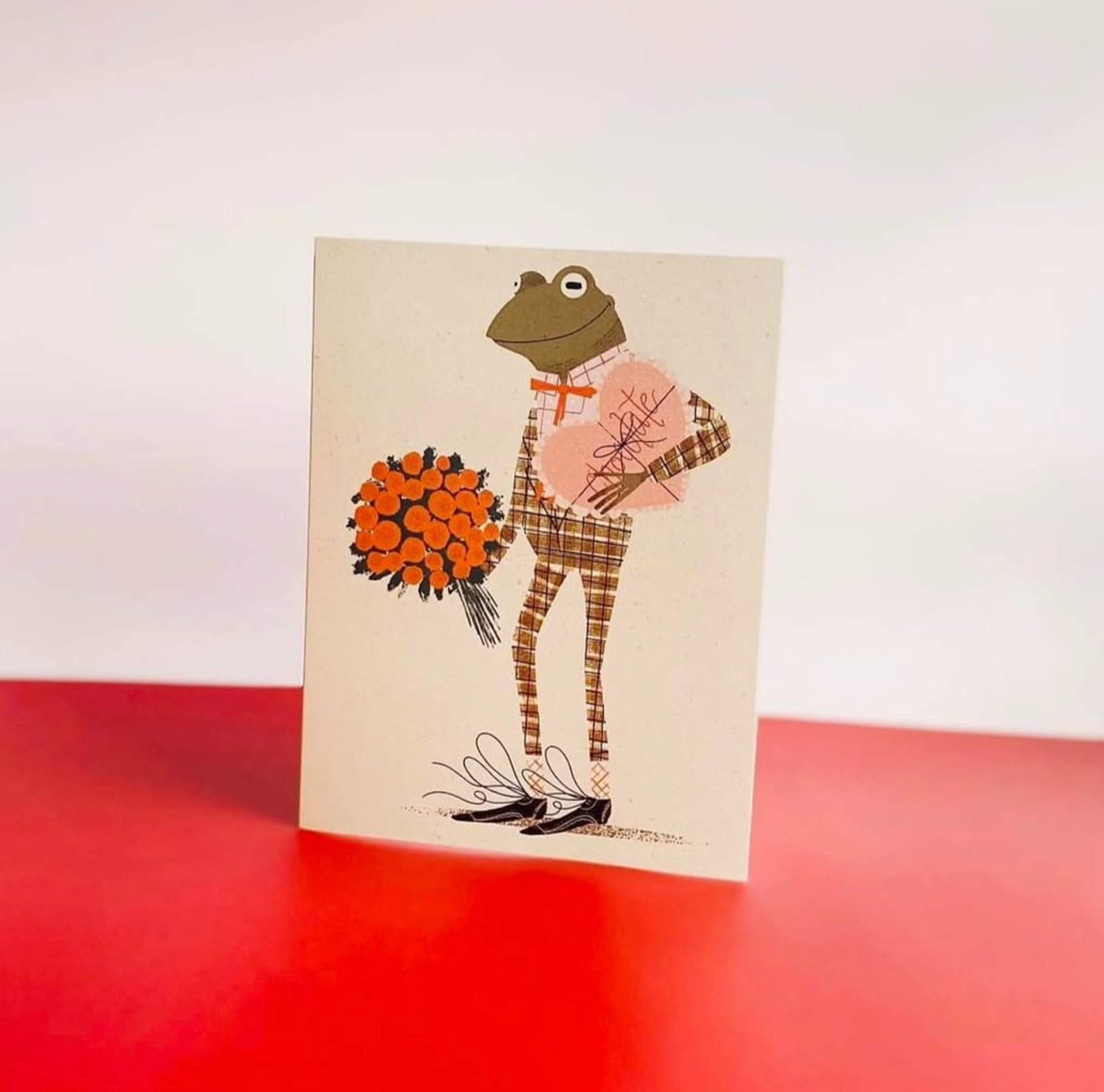 Greeting card with a stylized frog holding flowers on a red surface
