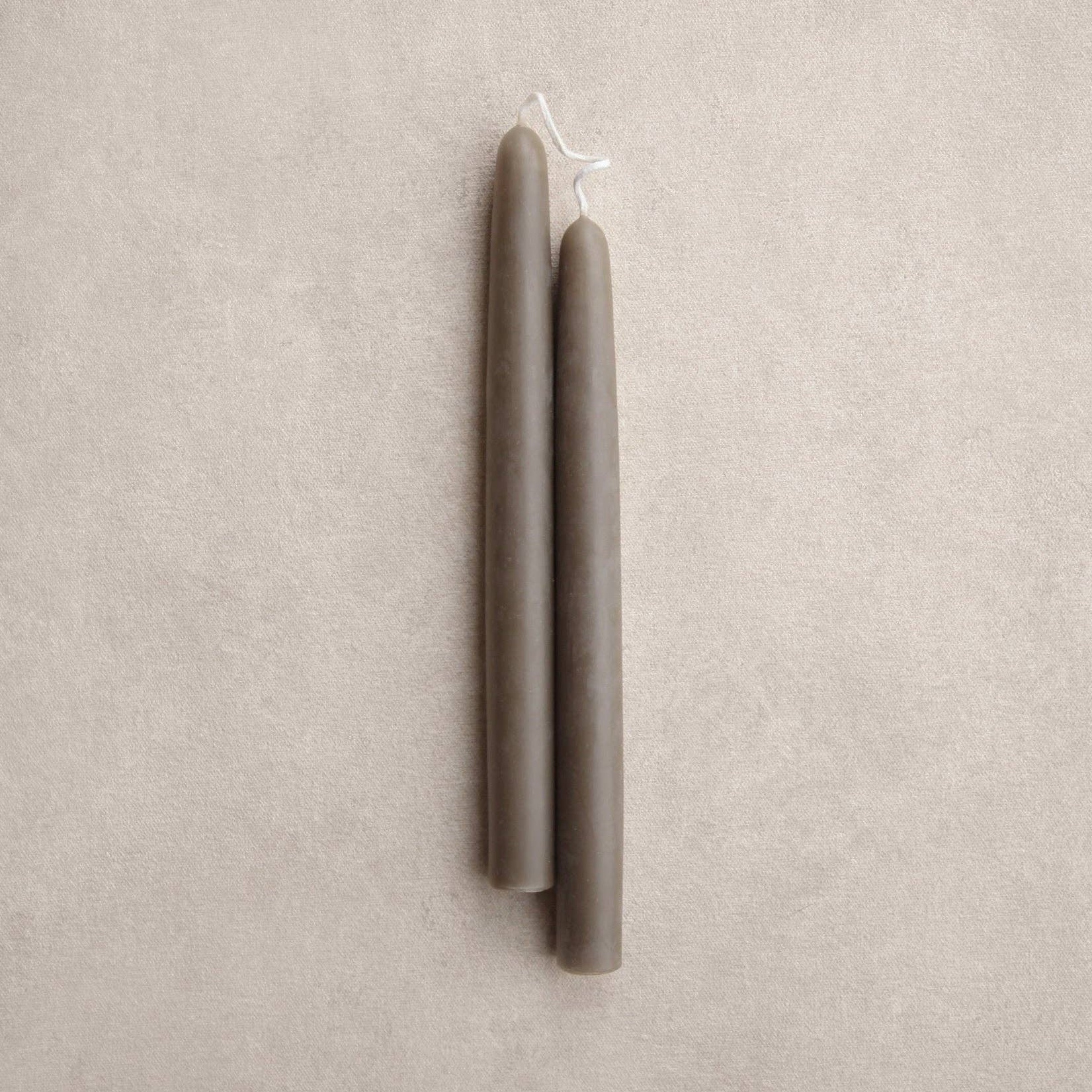 Two thyme colored taper candles standing upright side by side on a textured beige background.