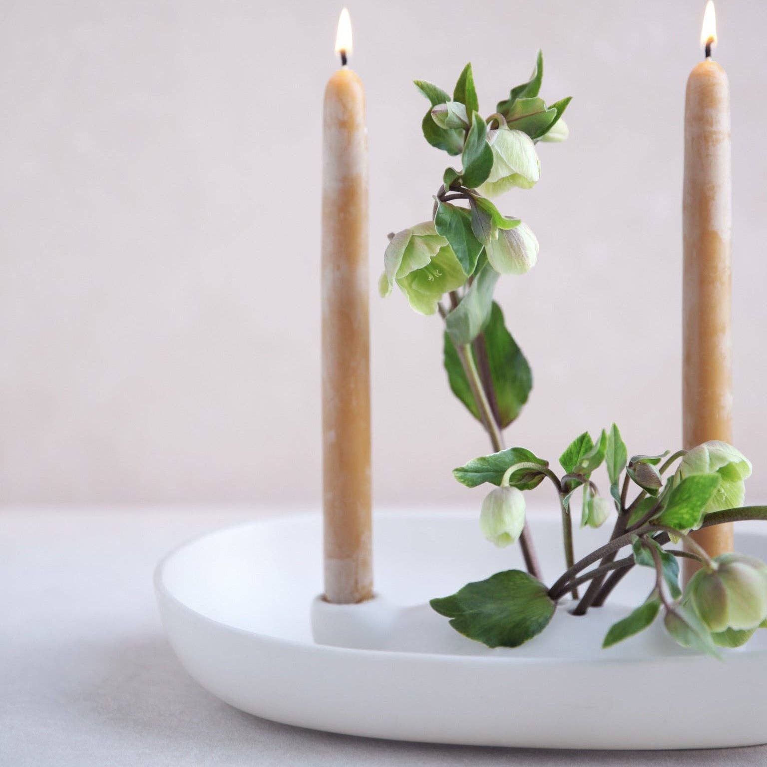 A white ceramic candle holder with two lit beige taper candles and green hollyhock branches, set against a soft, neutral background.