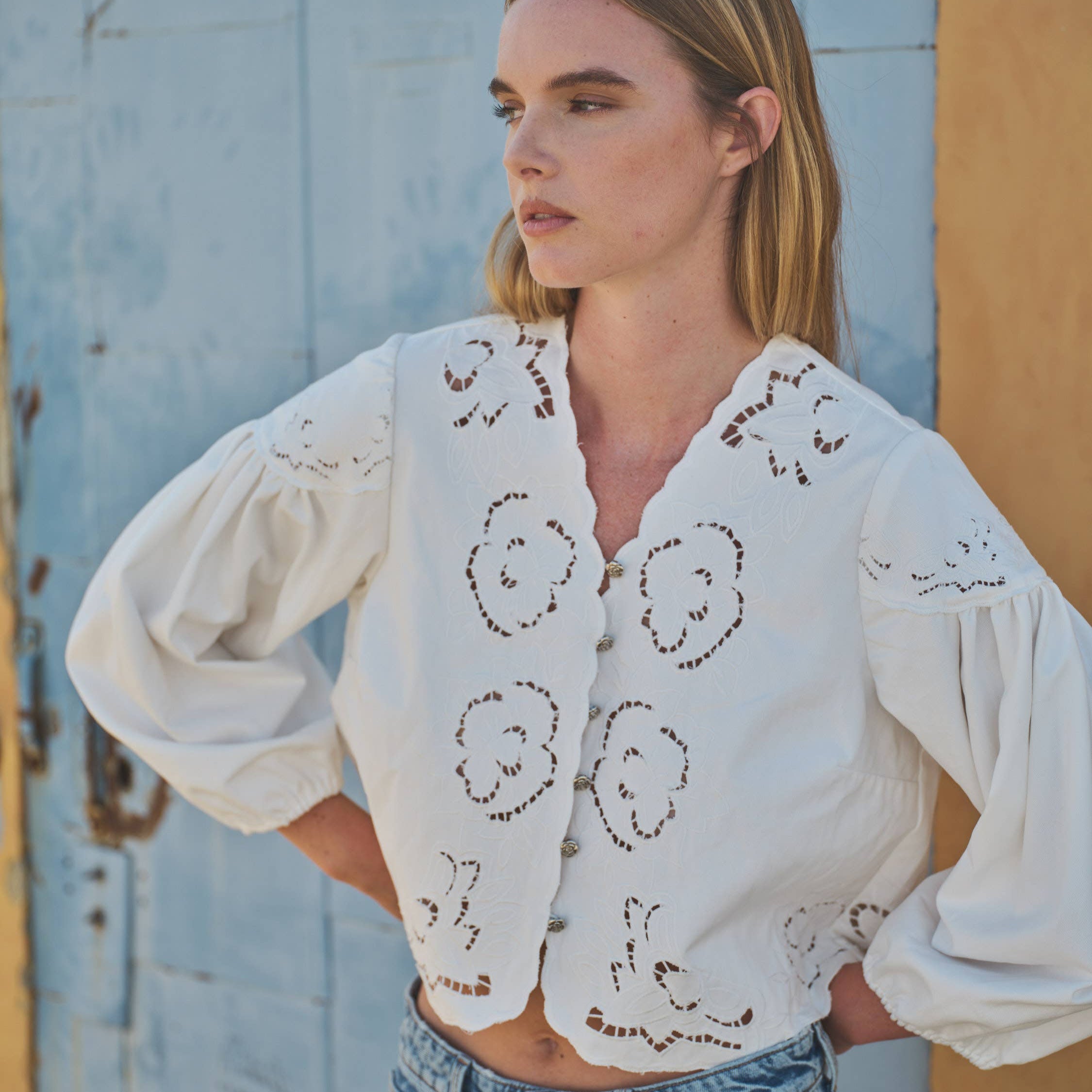 Woman wearing a white embroidered blouse and blue jeans against a rustic wall.