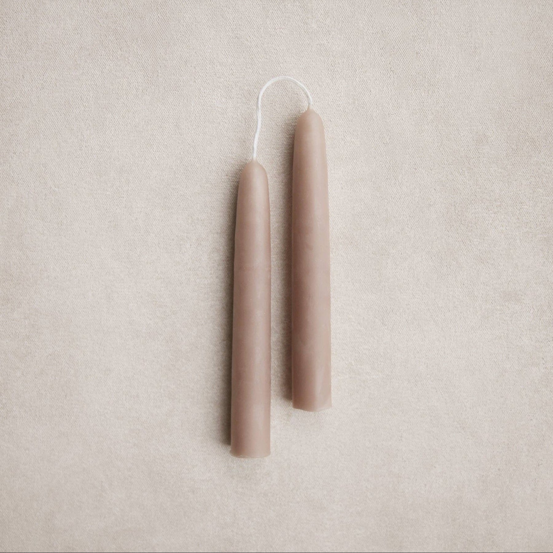 Two slim tapered candles in a muted mushroom color with white wicks, displayed vertically on a textured light beige background.