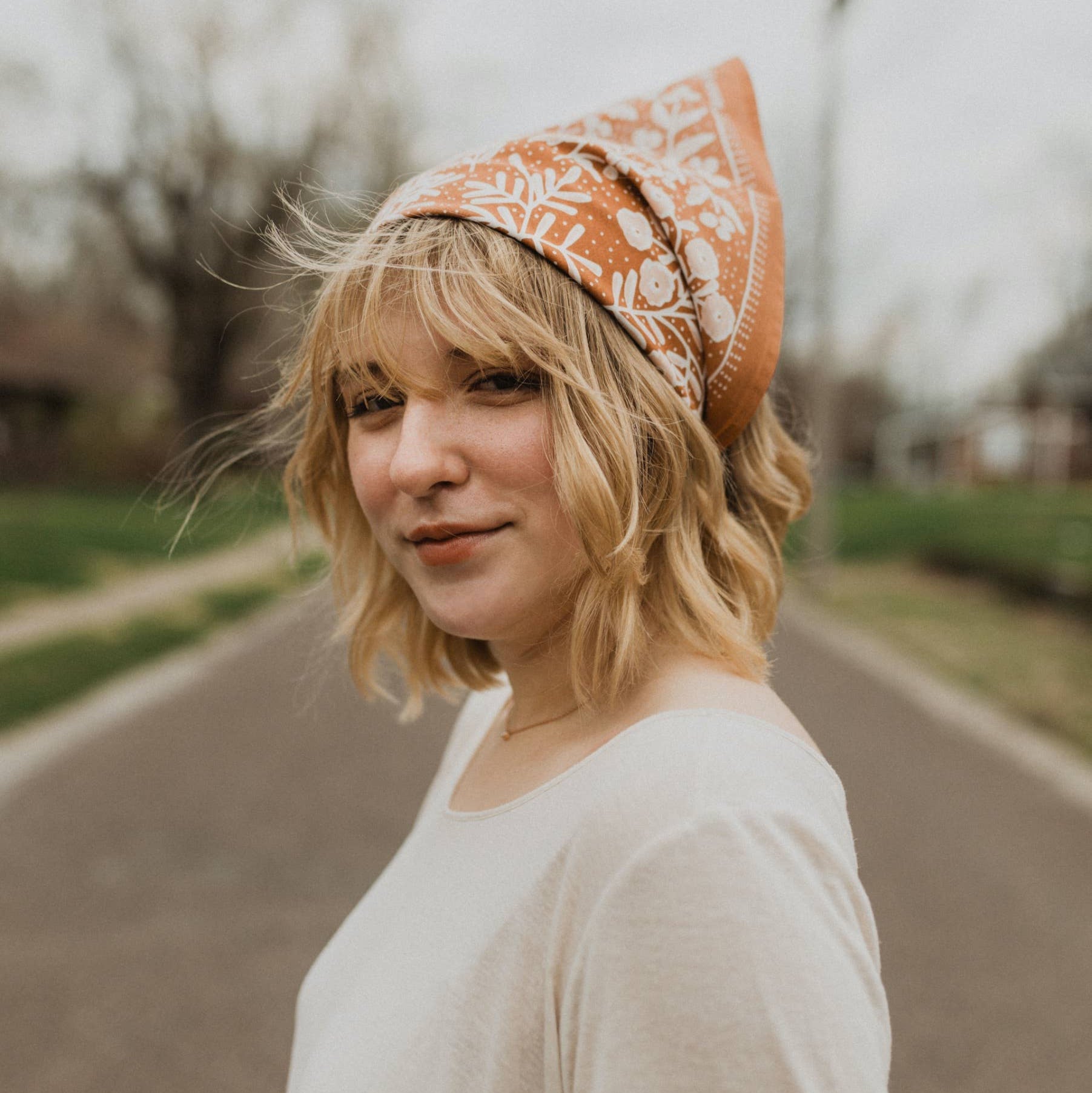 Woman wearing an orange bandana with a pattern, standing on a road.
