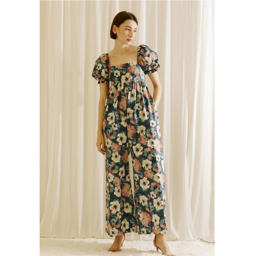 Large floral print baby doll jumpsuit. It exhibits a square neckline, side pockets, short puffy sleeves, and bust darts. It also has a gathered empire waist, wide palazzo legs, and an upper smocked back.