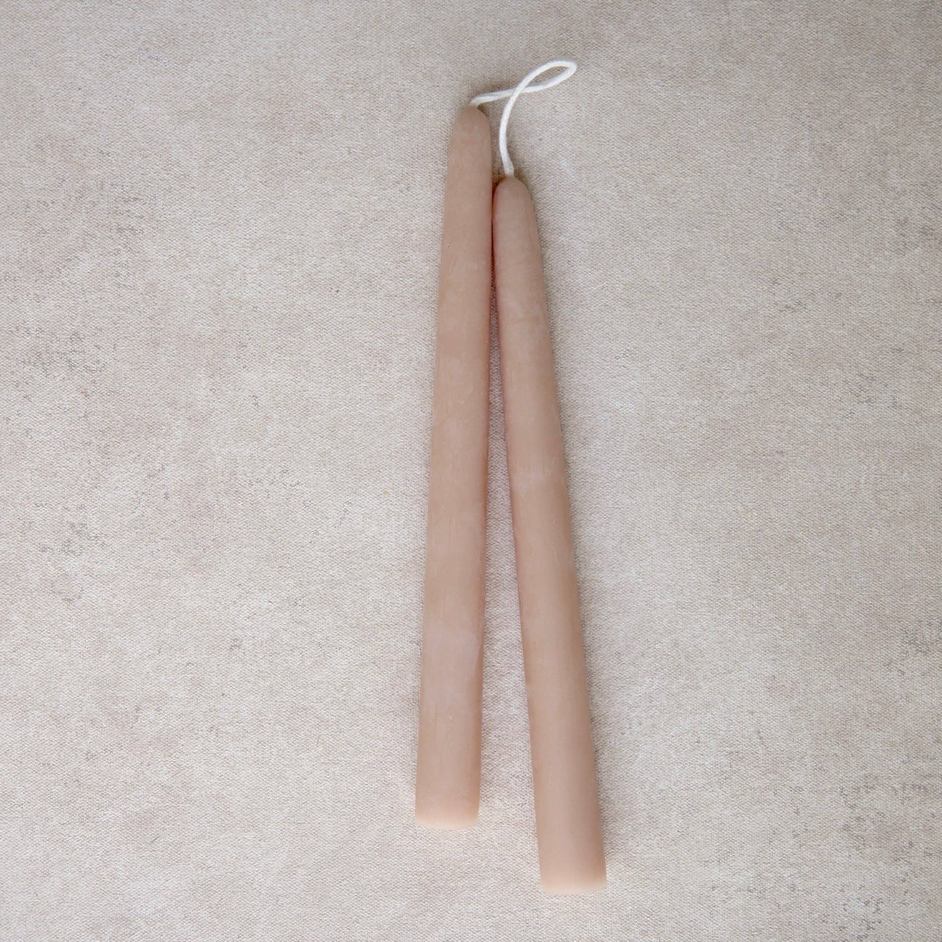 Two long taper candles in a light rose color, tied together with a white string at the top, displayed on a textured beige surface.