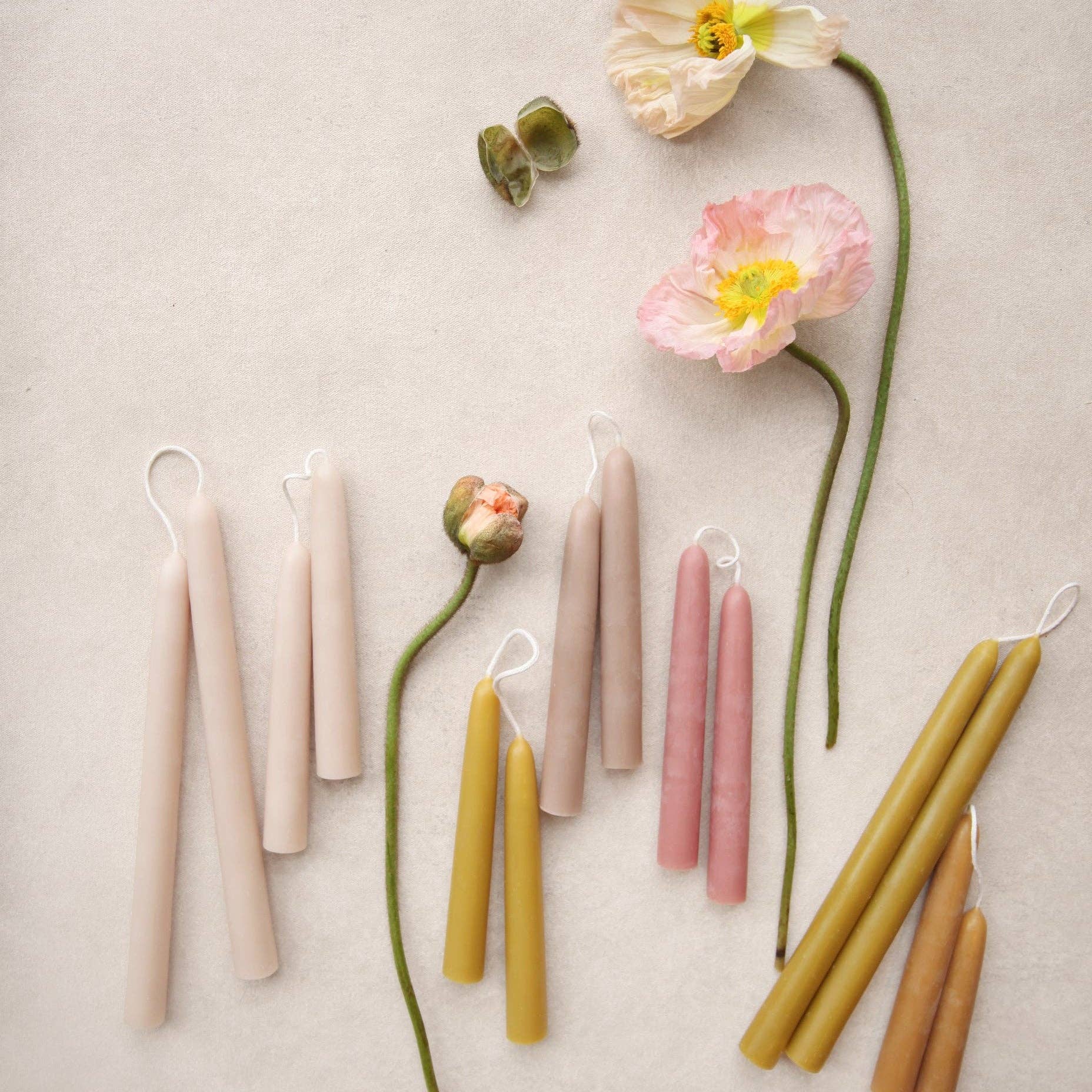 A styled flat-lay of multiple taper candles in soft pastel shades including blush pink, pale beige, mustard yellow, and warm tan, arranged on a light neutral background with decorative flowers and green stems.