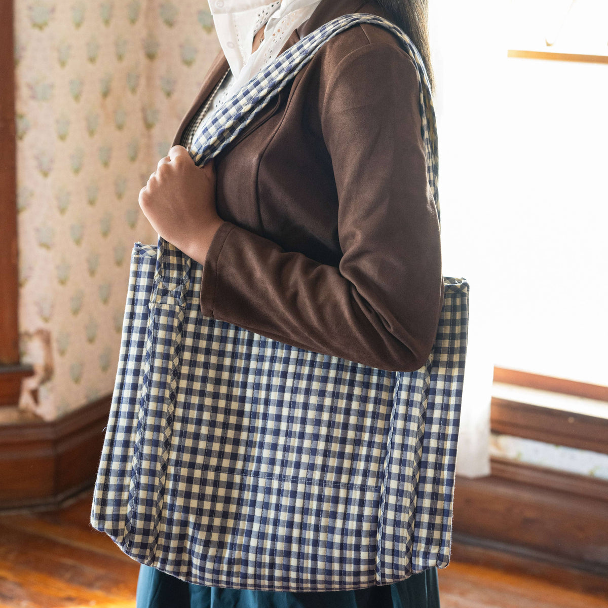 Woman wearing a checkered headscarf and apron in a room with patterned wallpaper.