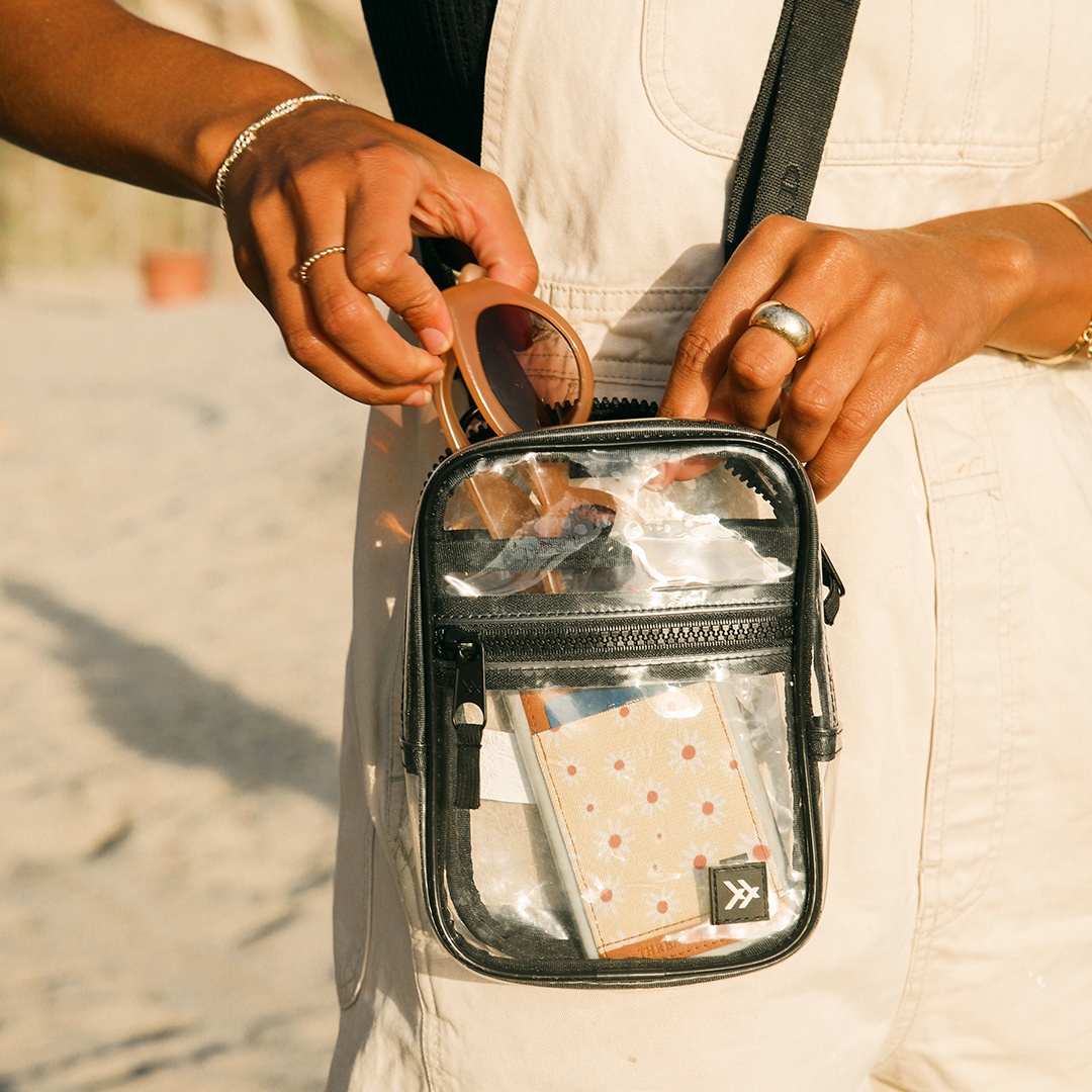 Person holding a clear bag with a black strap on a sandy background