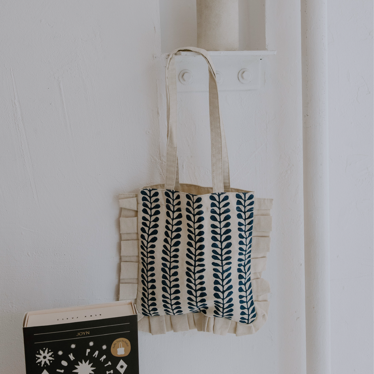 Tote bag with leaf pattern hanging on a white wall next to a book.