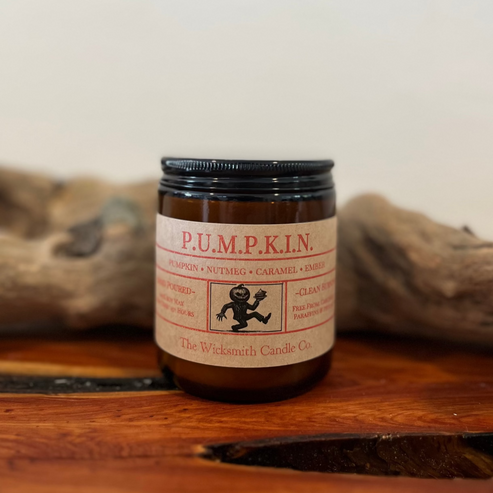 Amber glass candle jar labeled ‘P.U.M.P.K.I.N.’ by The Wicksmith Candle Co., featuring scent notes of pumpkin, nutmeg, caramel, and ember placed on a wooden surface with a piece of driftwood in the background