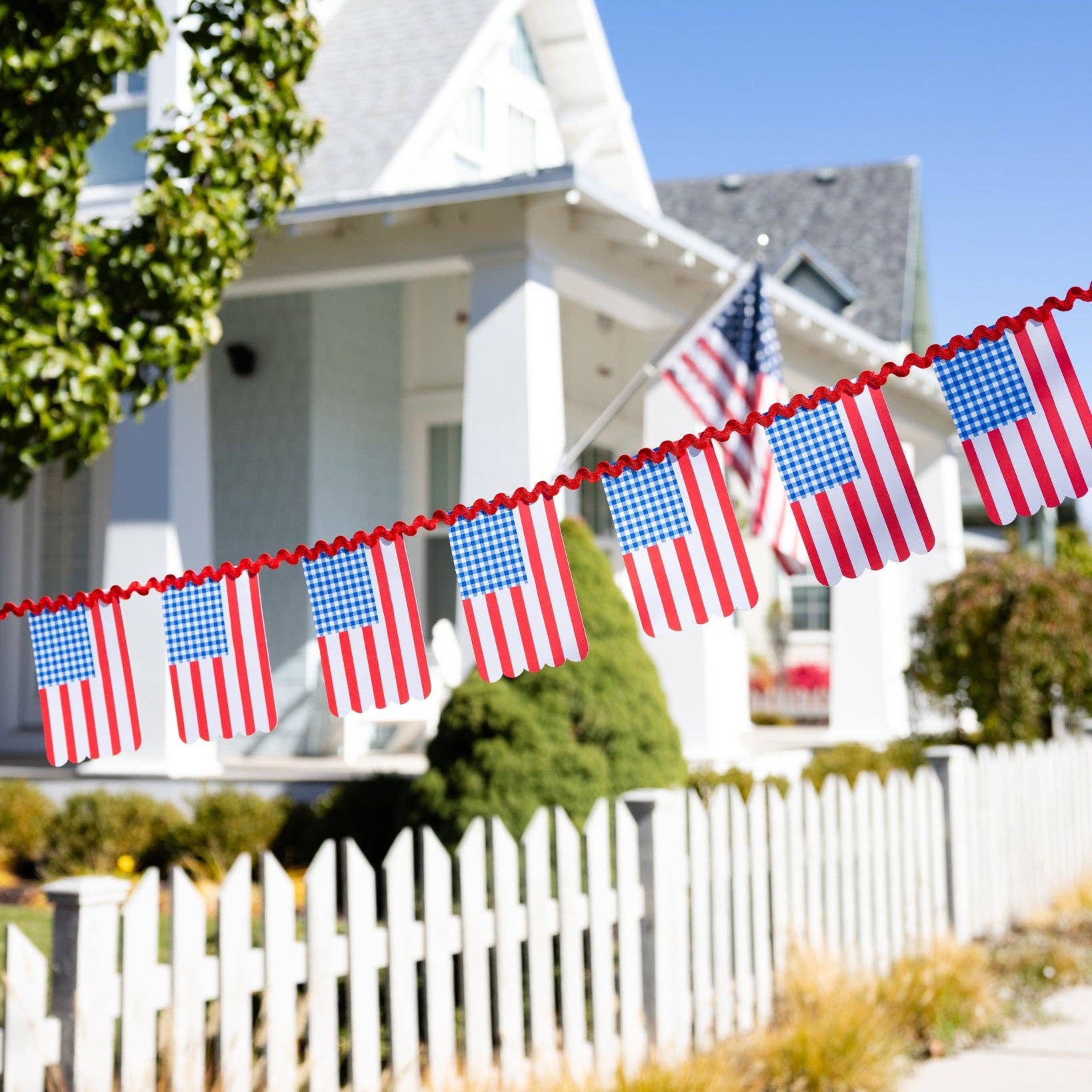 Celebrate in classic Americana style with our Gingham Flag Banner from the Floral and Flags Collection, a festive paper banner perfect for Fourth of July, Memorial Day, or any summer gathering. Featuring charming blue gingham flags with scalloped edges sewn onto a vibrant red ric-rac ribbon, this banner brings a cheerful, homespun touch to your patriotic celebrations.
