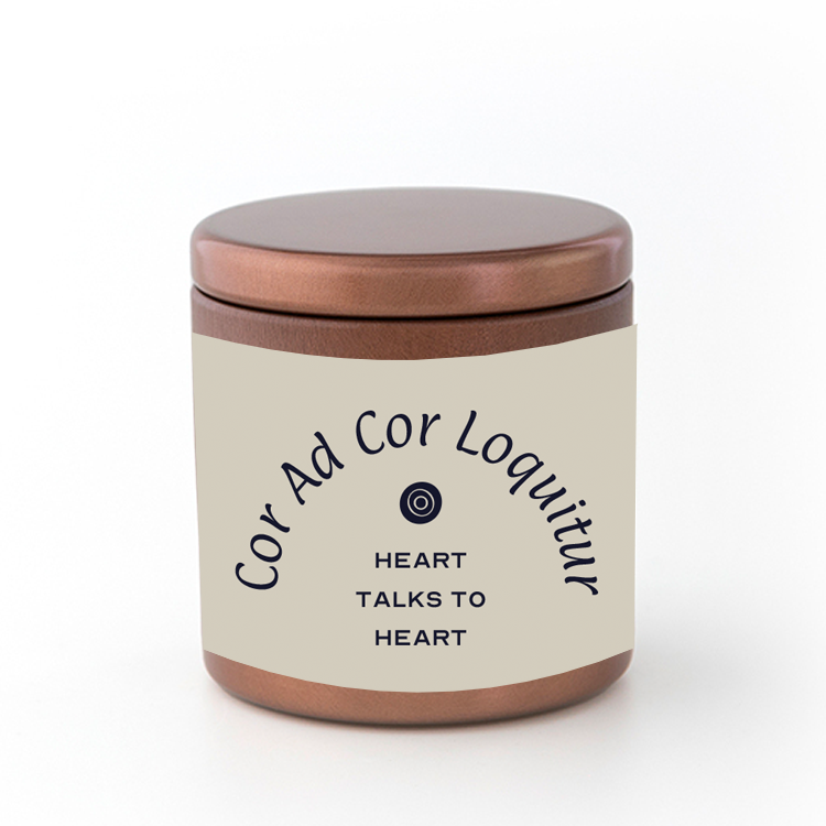 A bronze-colored travel candle in a metal tin with a label that reads 'Cor Ad Cor Loquitur - Heart Talks to Heart' on a white background.