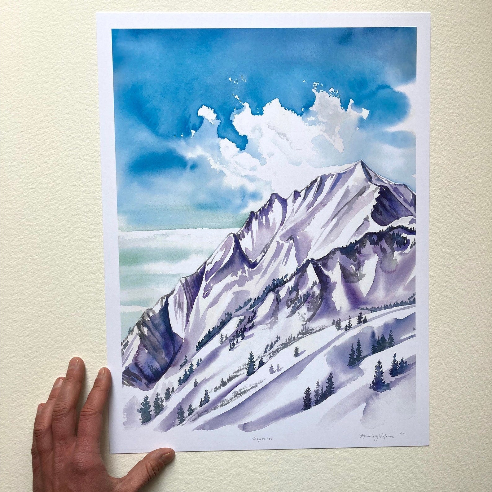Print against a white wall with a woman's hand. Painting one in blues and muted deep purples on the snowy mountain range. Pine trees dot the scape.