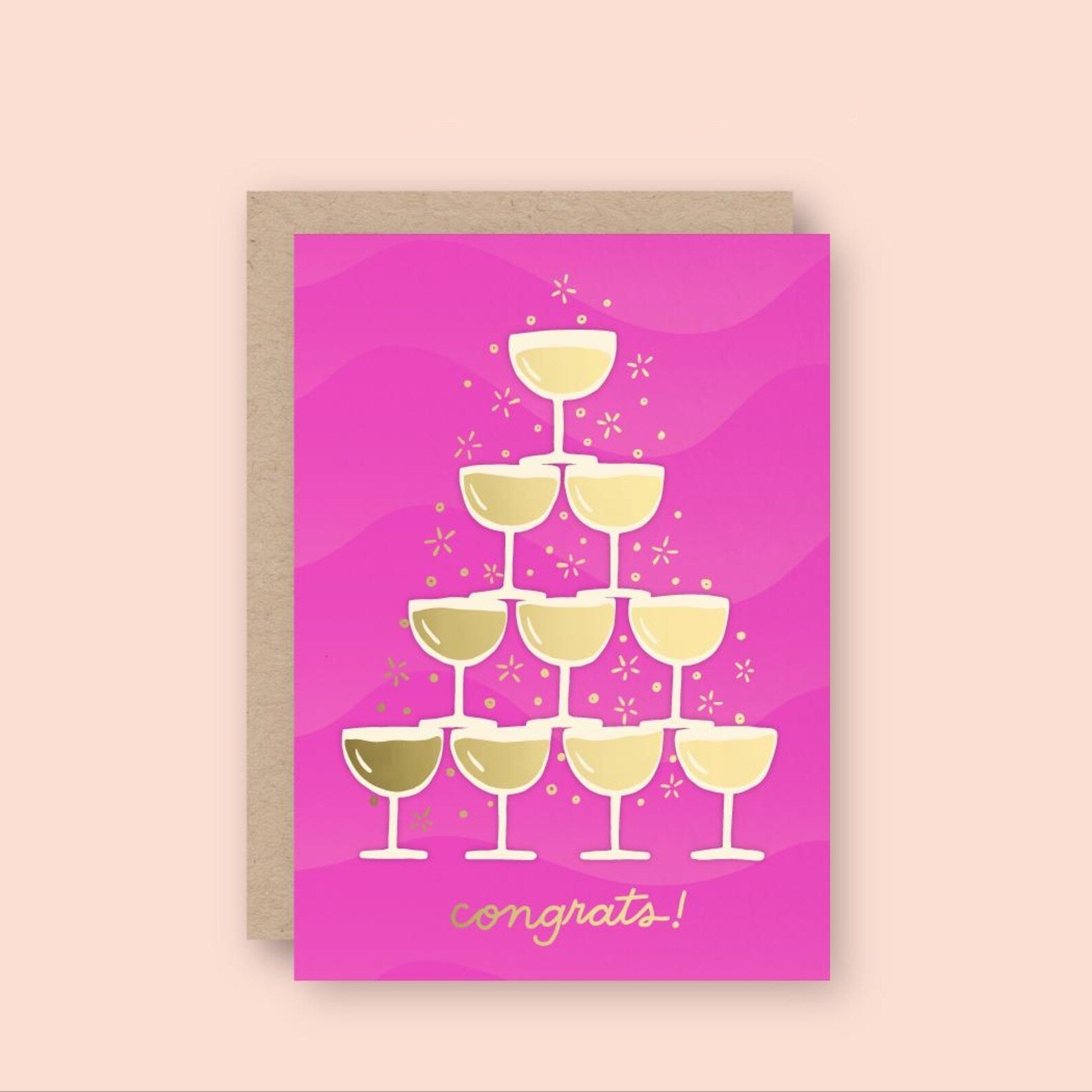 A bright pink greeting card features a gold foil illustration of a champagne tower made of ten glasses, with small celebratory sparkles around them. The word "congrats!" is written in shiny gold script at the bottom, and the card uses letterpress and foil stamping for a textured, festive finish.